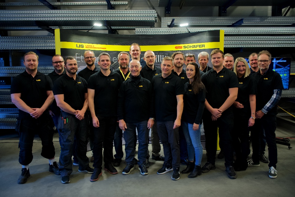 Et gruppebilde av tjue personer i matchende sorte polo-skjorter med firmaets logo, poserende smilende foran en gul og svart sikkerhetsbarriere i et industrielt lagermiljø.